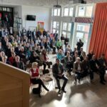 Besucher verfolgen die Eröffnung der PEGA Hausmesse 2024 während der offiziellen Begrüßung.